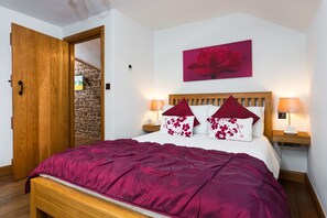 A bedroom with a wooden bed, a red floral pillow, a nightstand with a lamp, and a stone wall.