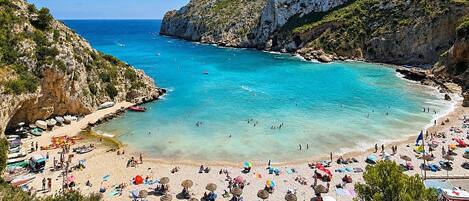 A beach with clear turquoise water, rocky cliffs, and a variety of beach umbrellas and sun loungers.