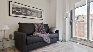 Un salon moderne avec un canapé noir, un tableau encadré représentant un paysage urbain, une table basse en verre, un lampadaire et une grande fenêtre donnant sur des immeubles.