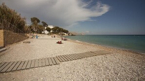 A pebble beach with a wooden walkway, palm trees, and a building in the background. - Finca Nora, Altea, Altea (Altea)