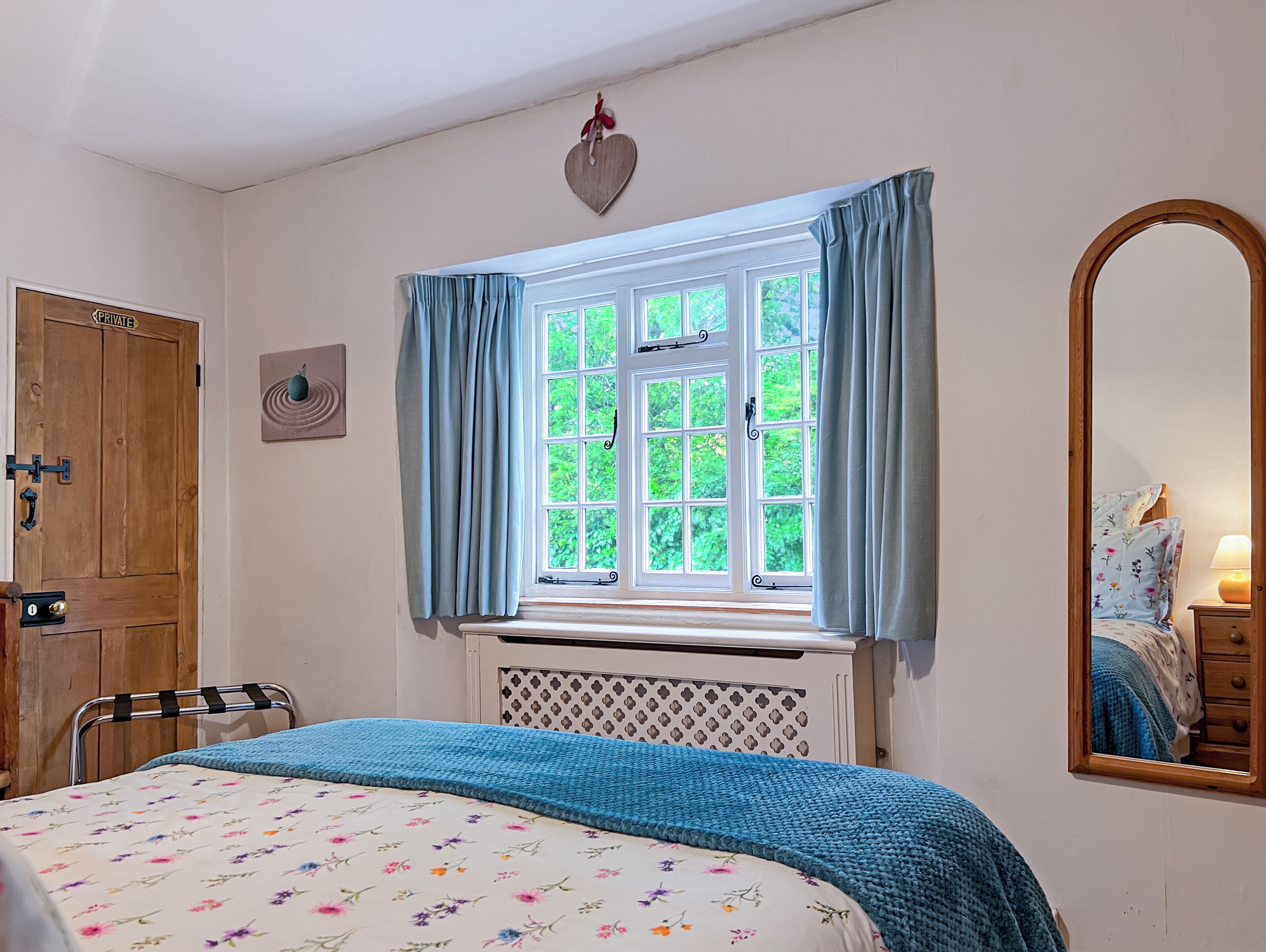 Quarto com cama, janela com cortinas, porta de madeira, espelho e aquecedor fixado na parede.