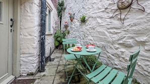A narrow outdoor patio with a table set for two, surrounded by green chairs, a white wall with a sun decoration, and potted plants.