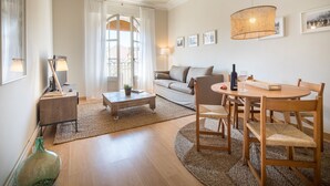 A modern living room with a round wooden dining table, a sofa, and a coffee table. There are framed pictures on the wall and a large window with curtains. - Bailen Terrace - Habitat Apartments (Barcelona)
