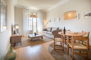 A modern living room with a sofa, coffee table, dining table, and chairs. There is a large window with curtains, a lamp, and framed pictures on the wall. - Sunny Bailen Flat with Private Terrace (Barcelona)