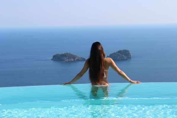 Une personne se tient au bord d’une piscine à débordement, avec une vue sur la mer et les îles.