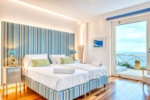 A double bed with white linens, a striped headboard, and a view of the sea through a large window.