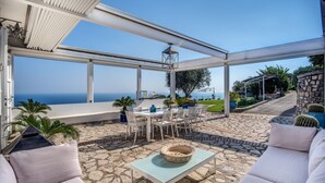 A covered outdoor patio with a stone floor, white furniture, and a view of the sea.