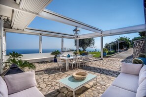 Une terrasse extérieure couverte, dotée d’un sol en pierre, de meubles blancs et offrant une vue sur la mer.