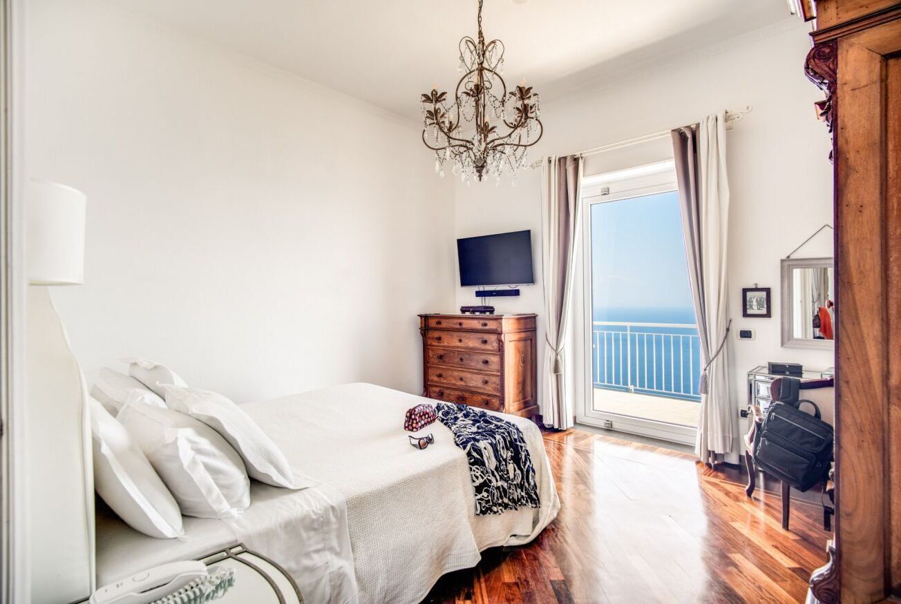 Une chambre à coucher comprenant un lit, une commode, un téléviseur, un balcon avec des rideaux et un lustre.