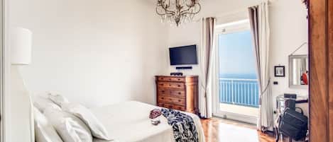 A bedroom with a bed, a dresser, a TV, a balcony with curtains, and a chandelier.