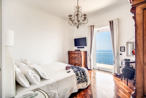 Ein Schlafzimmer mit Bett, Kommode, Fernseher, einem Balkon mit Vorhängen und einer Kristallleuchter.