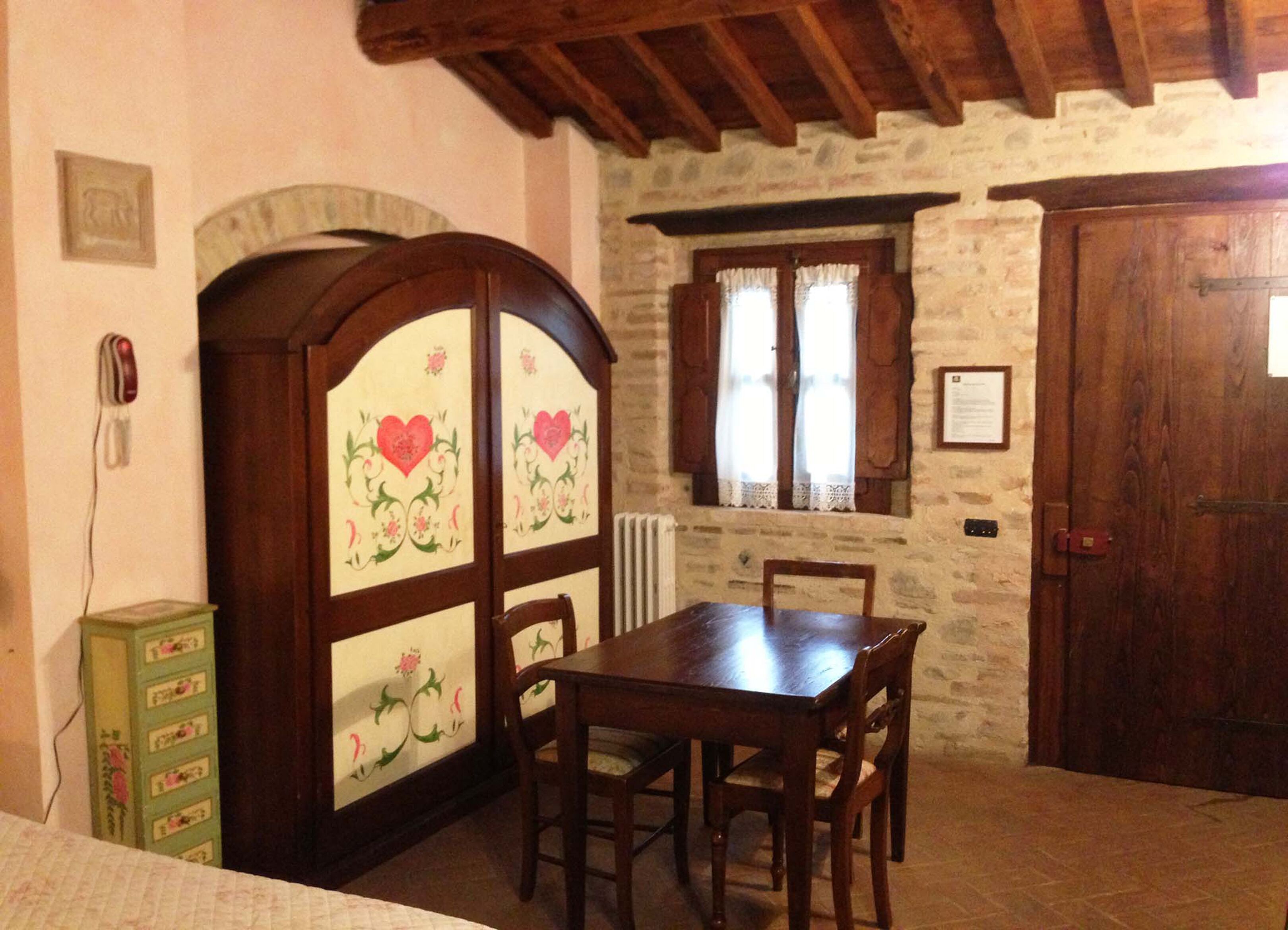 Habitació rústica amb taula de fusta, cadires, un armari amb motius florals i una paret de pedra.