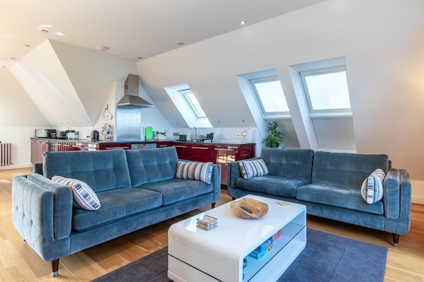 A modern loft apartment with two blue sofas, a white ottoman, and a kitchen in the background.