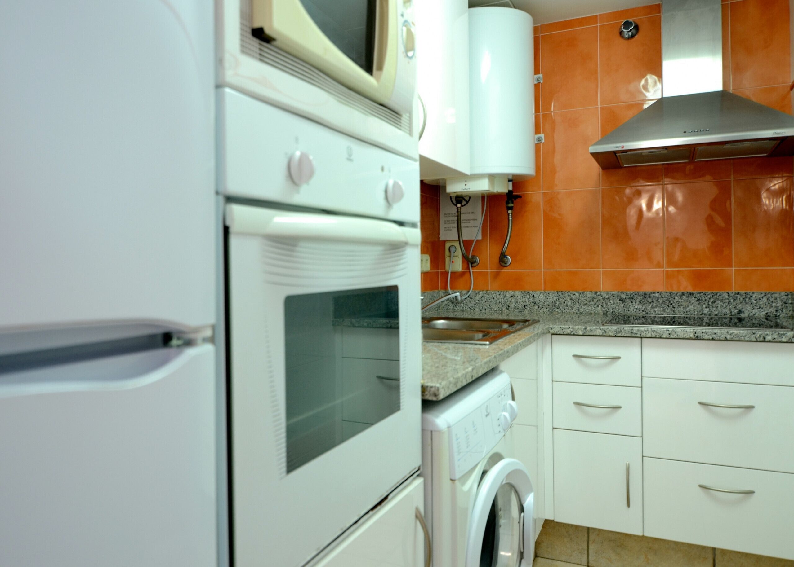 A kitchen with a white refrigerator, oven, and washing machine, a stainless steel range hood, and orange tiled backsplash.