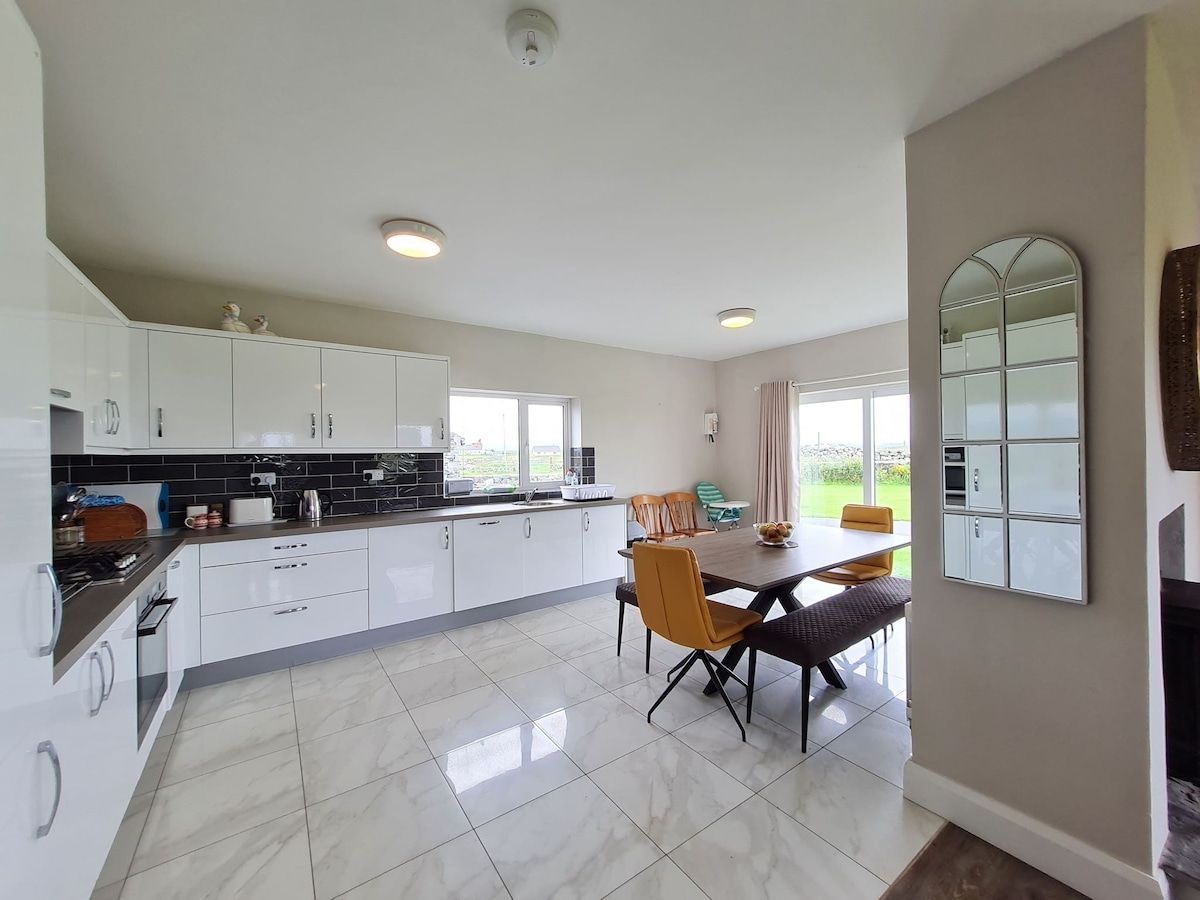A modern kitchen with white cabinets, a dining area with a table and chairs, and a large window providing a view of the outdoors.