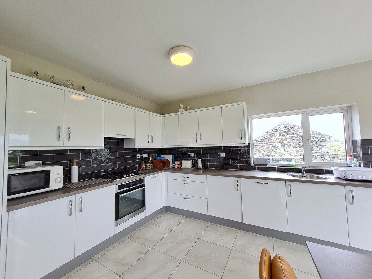 Une cuisine moderne avec des meubles blancs, un four à micro-ondes, une cuisinière et une fenêtre donnant sur une structure en pierre.