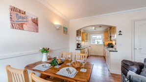 A dining area with a wooden table set for six, a kitchen in the background, and a beach-themed sign on the wall.