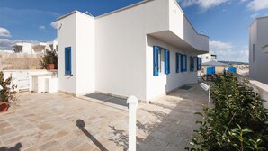 A modern white building with blue shutters, a stone-paved courtyard, and a view of the sea.