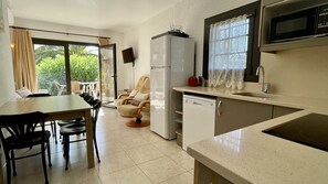 A compact kitchen with a refrigerator, microwave, and sink. There is a dining area with chairs and a table, and a chair with a cushion.