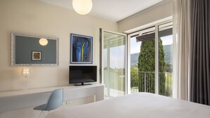A hotel room with a bed, a TV, a desk, a chair, a balcony with a view of greenery and hills. - Cà Gardesana With Pool (Garda)