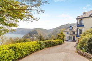 A coastal property with a paved driveway leading to a building with a balcony, surrounded by lush greenery and overlooking a body of water.