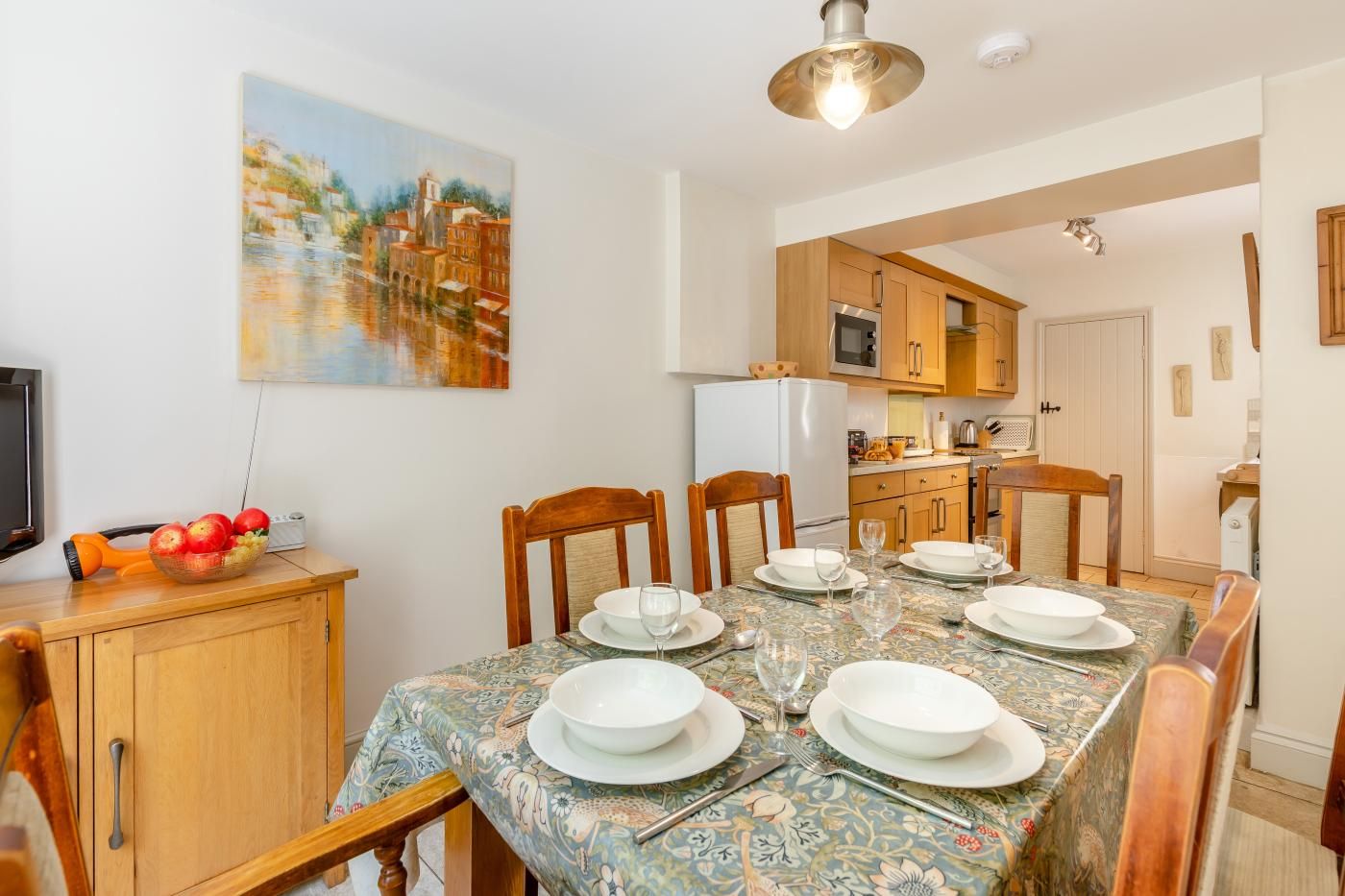 A dining area with a table set for four, wooden chairs, a painting on the wall, and a kitchen in the background.