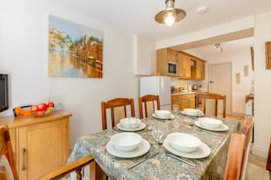 A dining area with a table set for four, wooden chairs, a painting on the wall, and a kitchen in the background.