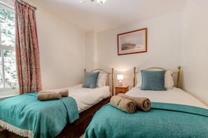 A room with two single beds, each with a headboard, a bedside table with a lamp, and a window with curtains.