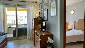 Ein Hotelzimmer mit einem Bett, einem Schreibtisch mit Computer, einem kleinen Tisch mit Lampe und einem Balkon mit Blick auf Gebäude.