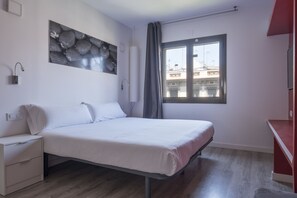 A modern bedroom with a bed, a nightstand, a wall-mounted lamp, a window with a view of buildings, and a red piece of furniture.