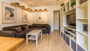 Een moderne woonkamer met een modulaire bank, een witte salontafel en een tv op een witte kast. Er hangen ingelijste foto's aan de muur en een plank met planten en boeken.