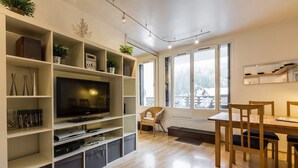 Een moderne woonkamer met een flatscreen televisie, inbouwkasten, een eethoek met een houten tafel en stoelen, en grote ramen met uitzicht op het landschap.