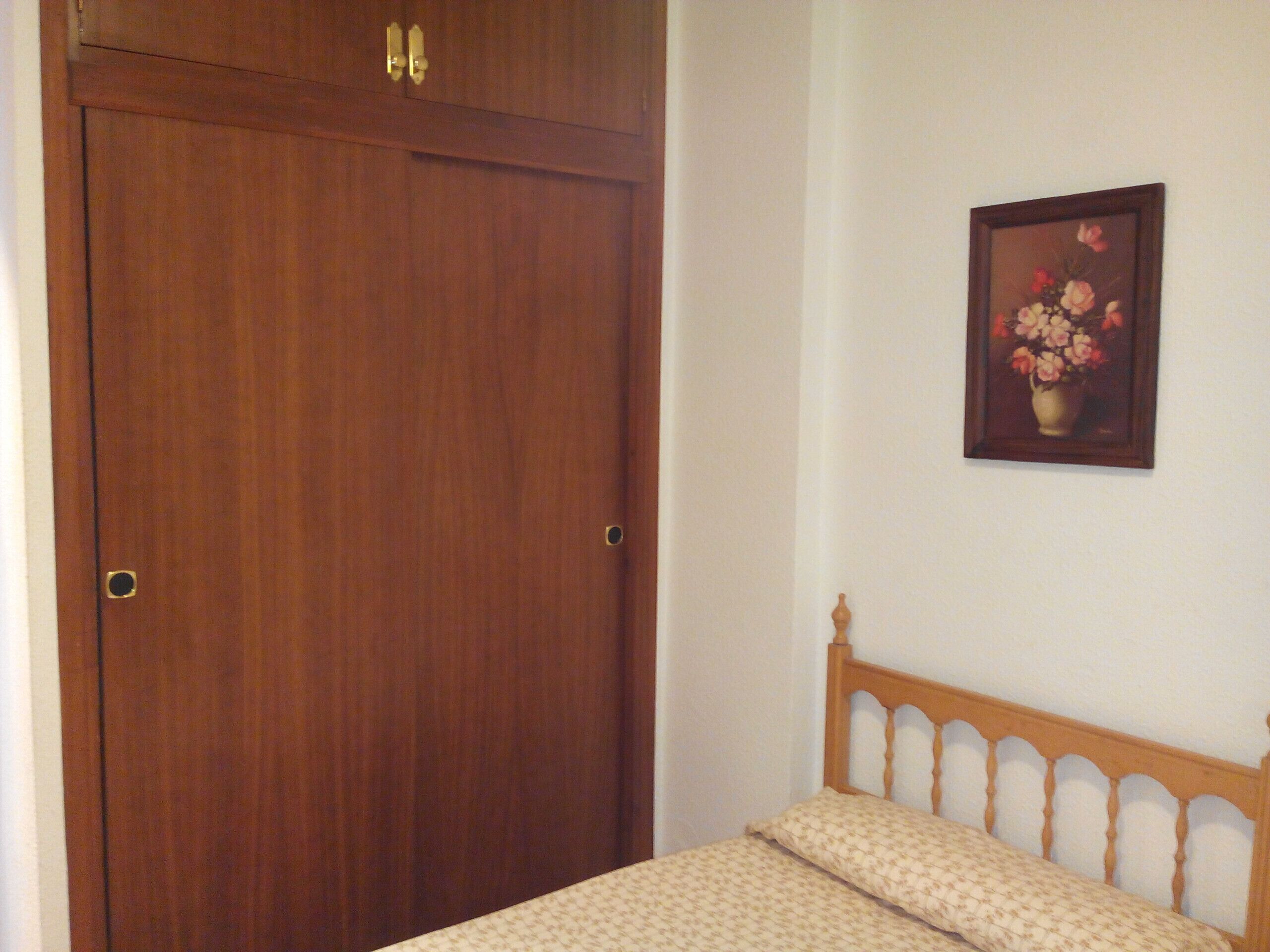 A bedroom with a wooden wardrobe, a bed with a wooden headboard, and a framed floral painting on the wall.
