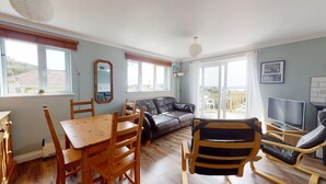 A living room with a sofa, a dining table with chairs, a TV, and a balcony with a view. - D59 Estuary View, Riviere Towans (Hayle)