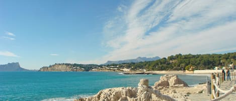 A coastal scene with a rocky shoreline, a wooden walkway, and a clear view of the sea and distant mountains.