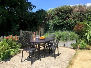 A round metal table with four chairs, a bottle of wine, and a bowl of fruit on a paved patio surrounded by lush greenery. - 2 Bed in Woolacombe (oc-high77) (Woolacombe)