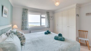 A bedroom with a bed, a chair, a window with curtains, and a view of the sea. - 2 Bed in Woolacombe (oc-high77) (Woolacombe)