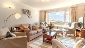 A well-lit living room with a sofa, armchairs, a coffee table, and a TV. There is a large window with a view of the hills. - 2 Bed in Woolacombe (oc-high77) (Woolacombe)
