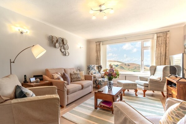 A well-lit living room with a sofa, armchairs, a coffee table, and a TV. There is a large window with a view of the hills.