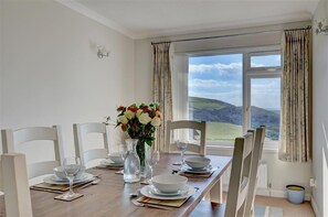 Ein Esszimmer mit einem Tisch für vier Personen, einer Blumenvase und Blick auf sanfte Hügel durch große Fenster.