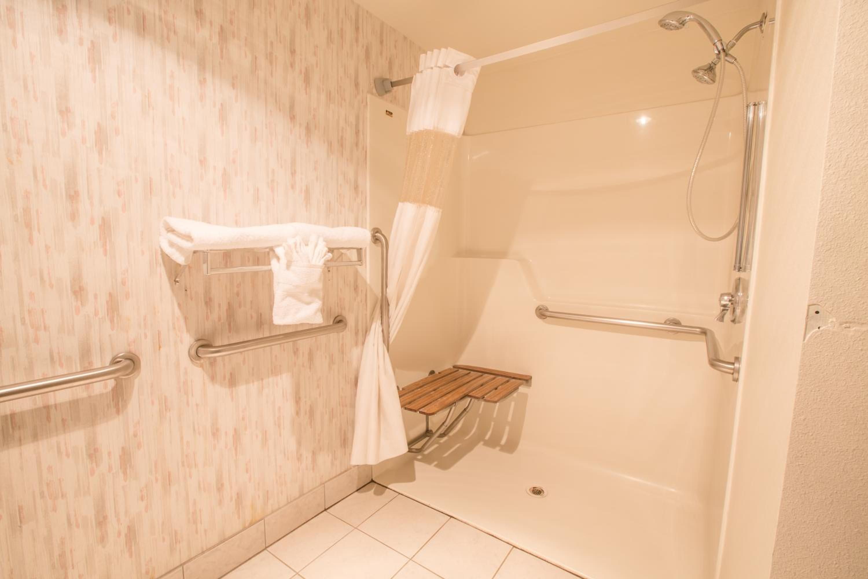 A bathroom with a shower, grab bars, a towel rack, and a small wooden shelf.