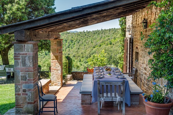 An outdoor dining area with stone pillars, a wooden pergola, and a view of a forested hillside.