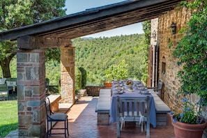 Un espace extérieur aménagé pour les repas, avec des piliers en pierre, une pergola en bois et une vue sur une colline boisée.