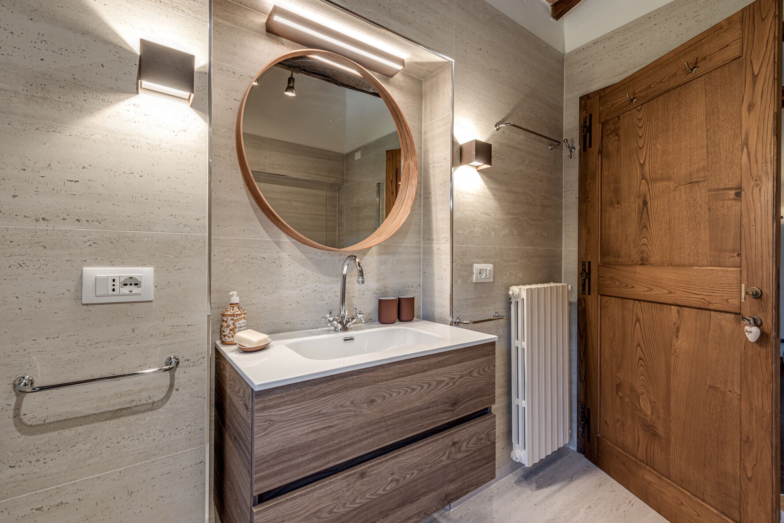 Un baño con tocador de madera, espejo redondo, lavabo blanco y puerta de madera.