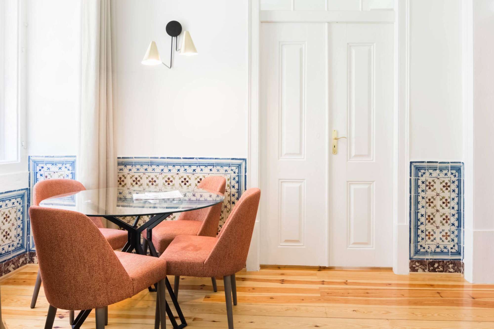 Un comedor con una mesa redonda, cuatro sillas, un aplique y una puerta con un borde de azulejos decorativos.