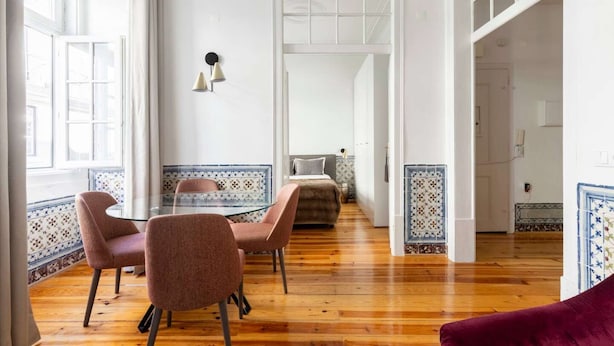 A spacious room with a wooden floor, a dining table with chairs, a sofa, and a bed with patterned tiles.