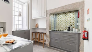 A modern kitchen with a marble countertop, grey cabinets, and a decorative backsplash. There are stools, a fire extinguisher, and a window with natural light.