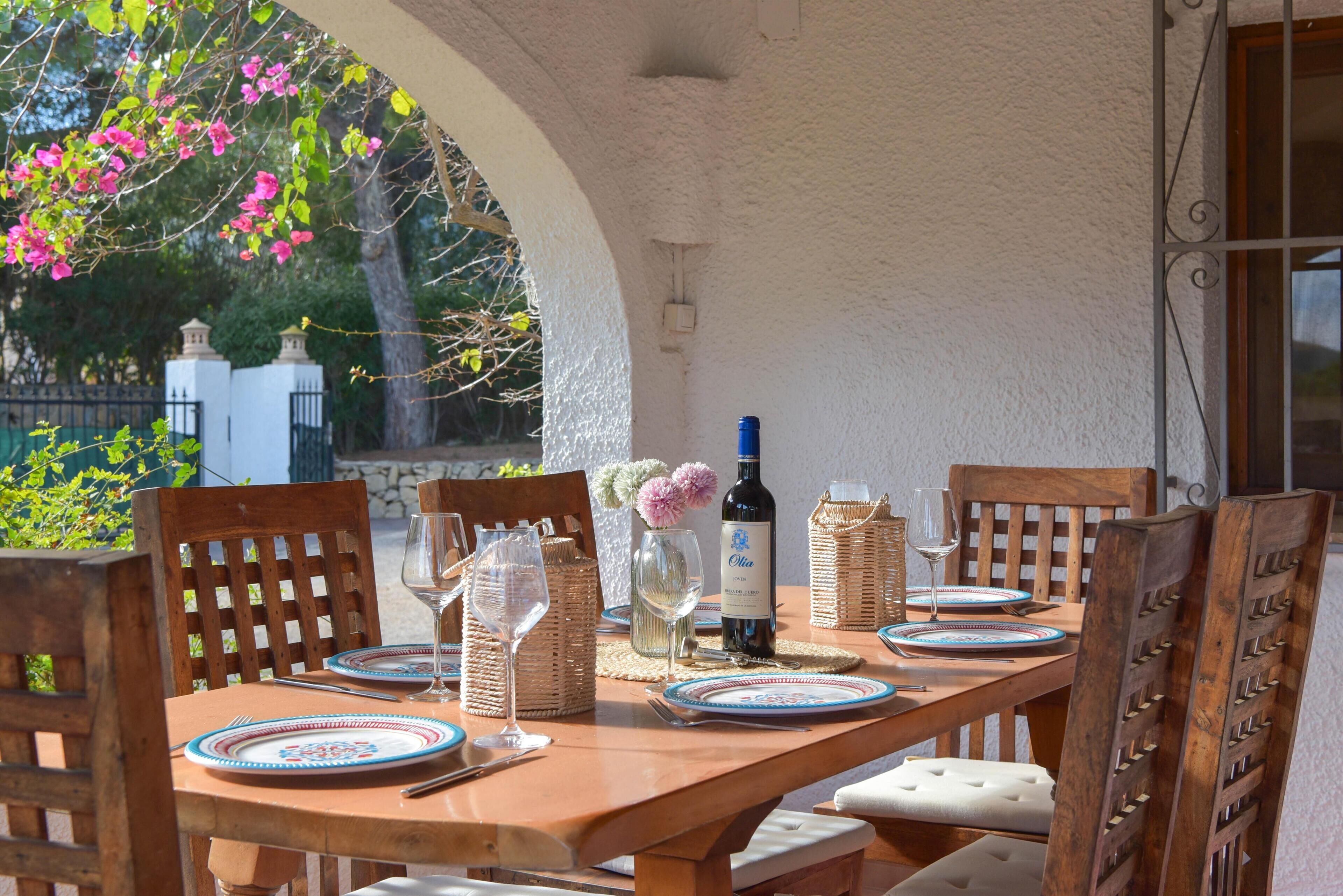 A wooden dining table set with plates, glasses, and a bottle of wine, surrounded by chairs with a white cushioned seat.