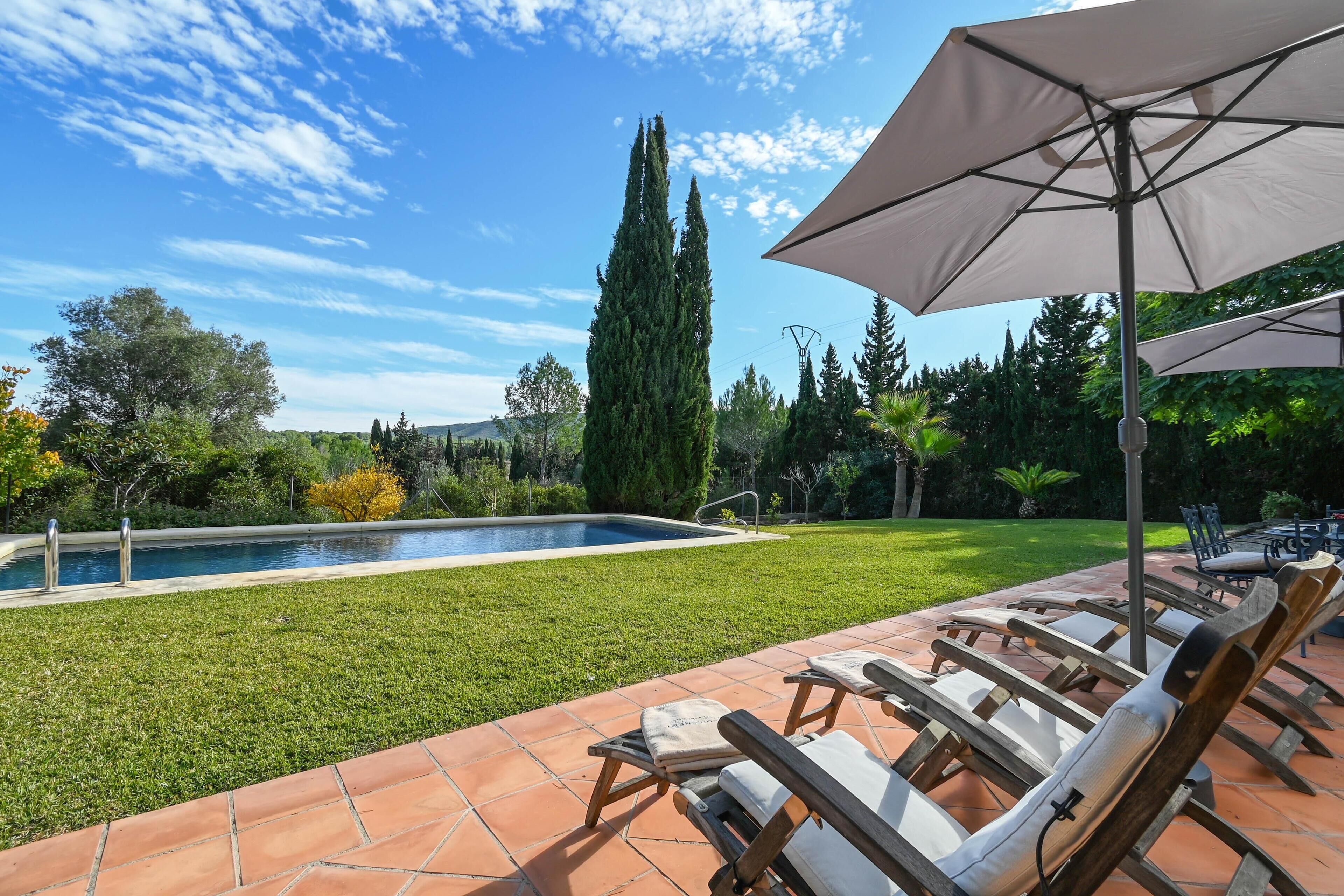 Un espace aménagé au bord de la piscine, avec des chaises longues et un grand parasol, entouré de verdure et surplombé par un ciel dégagé.