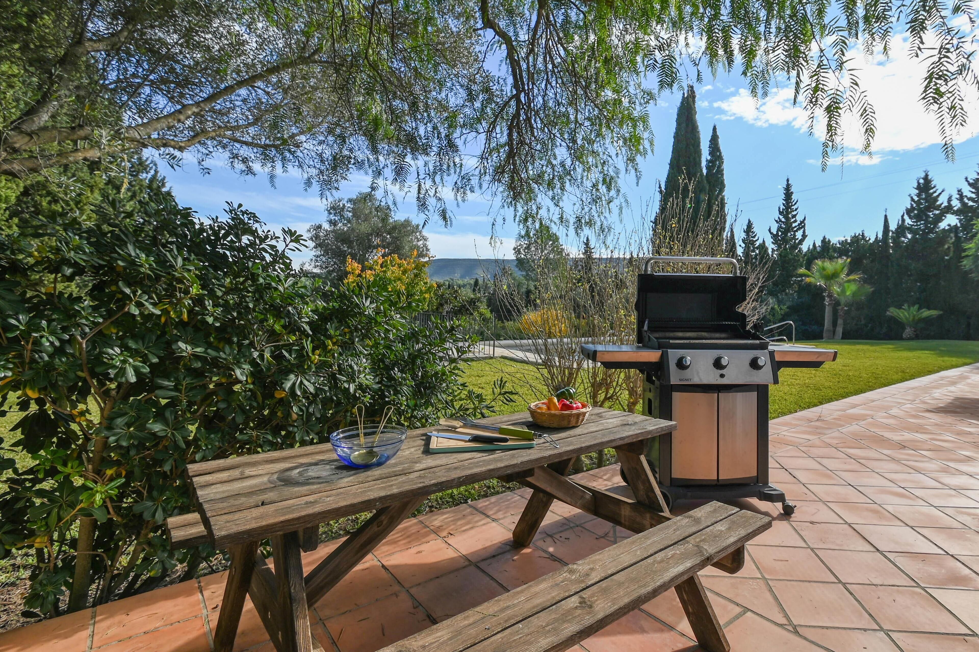 An outdoor patio with a wooden table and benches, a barbecue grill, and a view of a landscaped garden and distant hills.